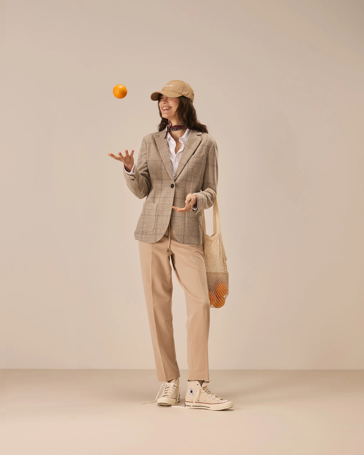 Vrouw in beige ruitjesblazer, beige pantalon en pet die een sinaasappel in de lucht gooit.