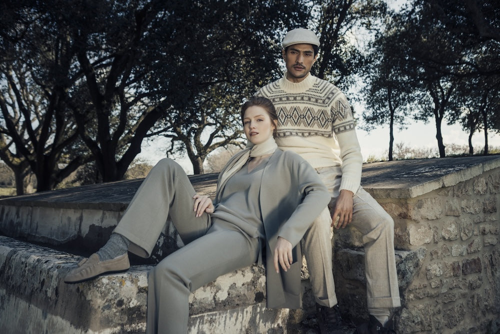 Donna con un completo grigio e uomo con un maglione chiaro a motivi, seduti su un muretto in pietra all'aperto.
